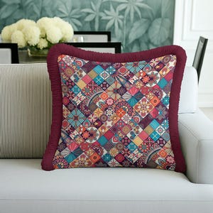 May include: Square decorative pillow with a colorful patchwork design. The pillow features a vibrant mix of geometric and floral patterns in shades of blue, orange, pink, and purple. It has a burgundy fringe trim and is placed on a white sofa.