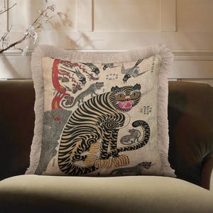 May include: A square throw pillow featuring a beige fabric cover with a fringe border. The print displays a large tiger with black stripes, a red mouth, and green eyes, surrounded by smaller tiger figures, birds, a red tree, and Japanese text.