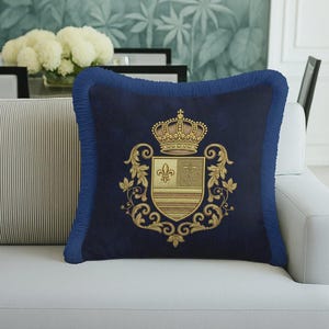 May include: Navy blue decorative pillow with a gold embroidered crest featuring a crown and fleur-de-lis. The pillow has a fringed border and is placed on a white sofa. The background includes a floral wallpaper.