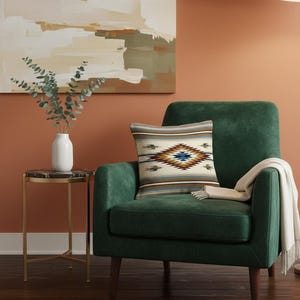 May include: A decorative pillow with a Southwestern design rests on a green armchair. The pillow features a central diamond pattern in shades of brown, blue, and cream, with striped borders. A white throw blanket is draped over the arm.