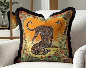 May include: Square decorative pillow with an orange background featuring two leopards. The pillow has a black fringe border and a detailed floral and animal print design. The pillow is placed on a white armchair.