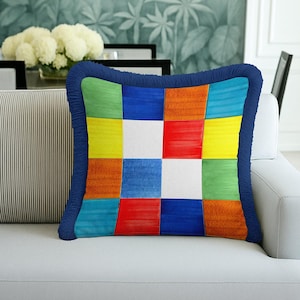 May include: Square throw pillow with a patchwork design in vibrant colors including blue, green, yellow, red, orange, and white. The pillow has a dark blue fringe border and is placed on a white sofa.