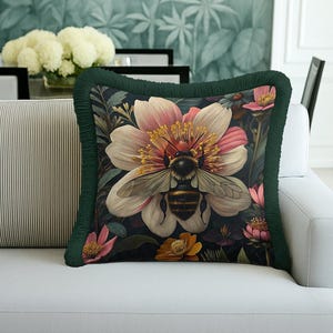 Puede incluir: Cojín cuadrado decorativo con una abeja en el centro de una flor con pétalos rosas y blancos. Tiene un borde con flecos verde oscuro. El cojín está sobre un sofá blanco, creando un ambiente floral.