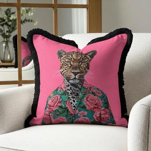 May include: A decorative pink throw pillow featuring a leopard wearing a floral shirt. The pillow has black fringe trim and is placed on a white armchair. The leopard's face is detailed with spots and a serious expression.
