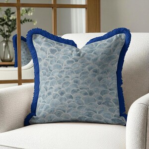 May include: A square decorative pillow with a blue seashell pattern and a dark blue fringe trim. The pillow is placed on a white armchair, creating a contrast of colors and textures. The pillow's design adds a touch of elegance to the room.