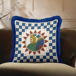 May include: Square decorative pillow with a blue fringe. The pillow features a white center with a colorful illustration of a person in a helmet surrounded by a red sunburst. The border has a blue and white checkered pattern.