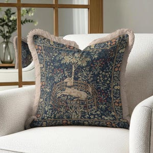 May include: Square cushion with a tapestry design depicting a unicorn in a garden. The cushion has a navy blue background with floral and tree details in beige, red, and green. It has a fringed edge and is placed on a white armchair. The cushion measures approximately 40 cm x 40 cm.