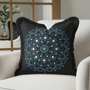 May include: Black decorative throw pillow with a blue and white mandala design. The pillow has a fringe trim and is placed on a white armchair. The mandala pattern is made of small dots.