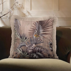 May include: Square decorative pillow with a botanical print featuring a bird, palm leaves, and flowers in shades of brown, gray, and green. The pillow has a fringed edge and is placed on a green sofa.