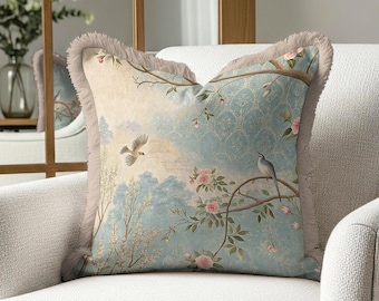 May include: Square cushion with a serene landscape design. The cushion features a light blue background with a bird, flowering branches, and a fringed edge. The cushion is placed on a white armchair.