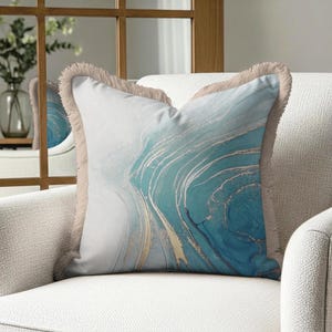 May include: A square throw pillow featuring an abstract marble-like pattern in shades of blue, white, and gold. The pillow is finished with a light beige fringe trim along all four edges, creating a textured border around the printed design.