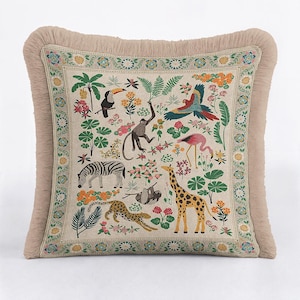 Puede incluir: Cojín cuadrado con flecos beige. El cojín presenta una colorida escena de la jungla con una cebra, una jirafa, un leopardo, un tucán, un mono y un loro. El diseño está rodeado por un borde floral.