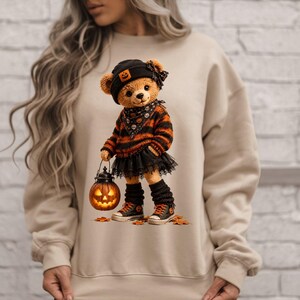 Puede incluir: Una sudadera beige de cuello redondo con el gráfico de un oso de peluche que lleva un gorro negro con un parche de calabaza, un suéter a rayas naranjas y negras, una falda de encaje negro, calentadores y zapatillas, sosteniendo una calabaza brillante.