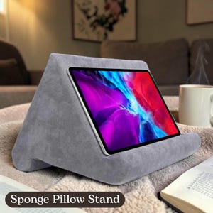 May include: A gray sponge pillow stand holding a tablet with a colorful screen. The stand is wedge-shaped with a curved base and the text "Sponge Pillow Stand" is at the bottom.
