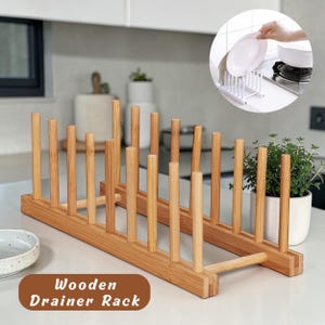 May include: A wooden plate drainer rack with vertical wooden dowels. The rack is made of light brown wood and has the words "Wooden Drainer Rack" in a brown box. A smaller white plastic plate rack is shown in the background.