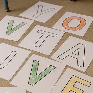Può includere: Biglietti di carta bianca con grandi lettere delineate che compongono la parola "VOTE" sono sparsi su un pavimento marrone chiaro. Le lettere sono colorate in verde, arancione e giallo. Le lettere sono delineate in nero.