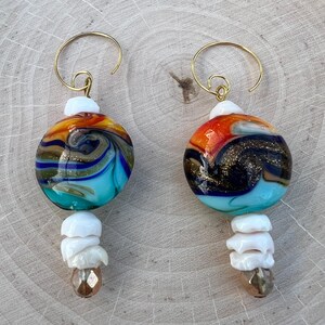 May include: A pair of earrings with a gold wire hoop and a round, multi-colored glass bead with white and gold accents. The beads are suspended from the hoops with white beads and a gold-toned bead.