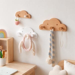 Puede incluir: Tres ganchos de pared con forma de nube hechos de madera y material pintado de blanco. Un gancho de madera natural sostiene una pequeña fresa de ganchillo, uno blanco sostiene una diadema de felpa y otro grande de madera sostiene una bufanda.