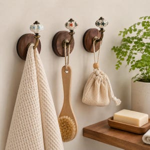 Peut inclure: Trois patères murales composées d'une base circulaire en bois foncé, d'un bras en métal doré et d'un bouton en céramique à motifs floraux. Elles supportent une serviette beige gaufrée, une brosse de bain en bois et un petit pochon.