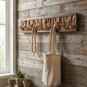 Puede incluir: Un perchero de pared de madera con un marco marrón oscuro y clavijas de madera natural. El perchero está adornado con un collar de cuentas y una bolsa tote beige. El fondo es una pared de madera rústica, con pequeñas plantas en macetas.