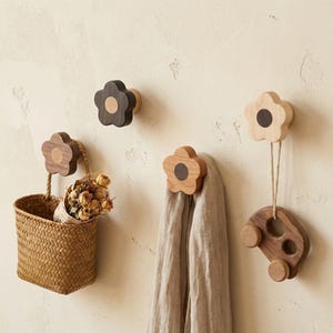 May include: A collection of flower-shaped wooden wall hooks in shades of brown and natural wood. One hook supports a woven basket with dried flowers, another a scarf, and a third a wooden car toy. The hooks are mounted on a beige wall.