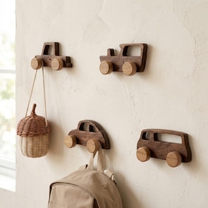 May include: Four wooden car-shaped wall hooks in different designs. Each hook has a wooden peg for hanging items. A woven basket and a backpack hang from two of the hooks. The hooks are mounted on a light-coloured wall.