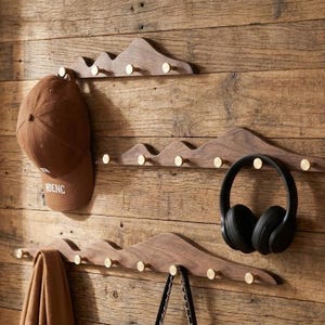 Puede incluir: Tres percheros de pared de madera con forma de montaña y pomos de latón están montados en una pared de madera. Una gorra de béisbol marrón, unos auriculares negros, una bufanda marrón y un bolso negro cuelgan de los ganchos. La pared tiene vetas de madera natural.
