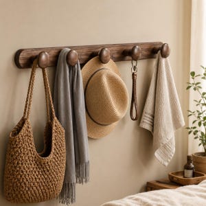 Könnte beinhalten: Eine horizontale Wandgarderobe aus dunkelbraunem Holz mit fünf abgerundeten Haken. An den Haken hängen eine beige gewebte Tragetasche, ein grauer Schal mit Fransen, ein beiger Strohhut, ein brauner Schlüsselanhänger aus Leder und ein beiges Handtuch.