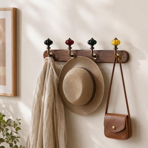 May include: A wall-mounted wooden coat rack featuring four brass hooks topped with decorative pumpkin-shaped knobs in black, red, dark green, and yellow. A beige fabric garment, a straw hat, and a brown leather shoulder bag hang from the hooks.