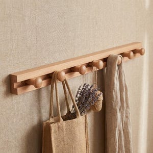May include: A wooden coat rack with eight rounded pegs. A beige tote bag, a bundle of lavender, and a light-colored scarf hang from the pegs. The rack is mounted on a neutral-toned wall.