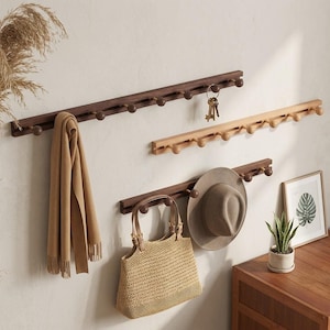 Könnte beinhalten: Drei hölzerne Garderobenleisten mit abgerundeten Haken sind an einer hellen Wand montiert. Ein Schal, Schlüssel, ein Hut und eine Handtasche hängen an den Leisten. Die Leisten sind aus dunklem und hellem Holz gefertigt.