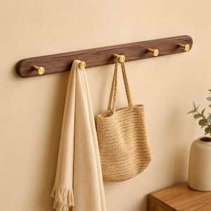 Peut inclure: Un porte-manteau mural horizontal en bois brun foncé doté de cinq crochets cylindriques en laiton. Un tissu texturé de couleur crème est suspendu au premier crochet et un sac fourre-tout en paille beige est accroché au deuxième crochet.