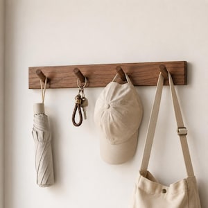 May include: A rectangular dark wood wall-mounted coat rack featuring four cylindrical wooden pegs. Hanging from the pegs are a folded beige umbrella, a set of metal keys on a braided brown leather keychain, a beige baseball cap, and a beige canvas bag.