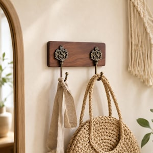 May include: A rectangular dark brown wooden wall-mounted rack featuring two ornate antique brass-toned metal hooks. The hooks have intricate decorative tops and curved ends. A beige linen fabric strip hangs from one hook and a woven straw bag hangs from the other.
