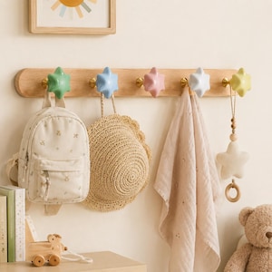 Puede incluir: Un perchero de madera clara con cinco ganchos en forma de estrella en colores pastel: verde, azul, rosa, blanco y amarillo. Los ganchos están montados sobre una tabla horizontal. Incluye una mochila pequeña, un sombrero de paja y una tela.