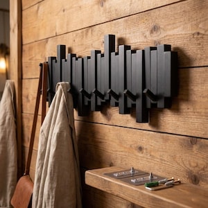 May include: A black wooden coat rack with multiple hooks is mounted on a wooden wall. The rack has a modern design with vertical rectangular elements. A tan jacket and a brown leather bag hang from the hooks. A small wooden shelf is below the rack.