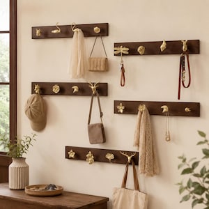 May include: Five horizontal dark brown wooden wall-mounted racks featuring various gold-toned metal hooks shaped like animals, flowers, and leaves. The hooks hold items including a beige scarf, a small handbag, a baseball cap, and a leather leash.