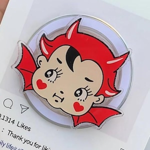 May include: A clear acrylic pin with a cartoon devil design, featuring red horns, wings, and heart-shaped cheeks. The pin is set against a mock Instagram post with the text "Album" and "Instagram".