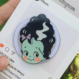 May include: A round, cartoon-style pin featuring a green-skinned character with black hair, a pink bow, and rosy cheeks. The pin is set against a white background with an Instagram-themed frame, including the word "Instagram" and a heart icon.