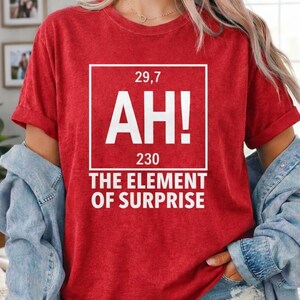 May include: Red t-shirt with a white graphic that reads "AH!" with the numbers 29,7 and 230 above and below the text. The text "THE ELEMENT OF SURPRISE" is below the graphic. The shirt has short sleeves.