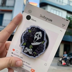 以下が含まれることがあります： 白黒のスクリームをテーマにした電話グリップ。スカルとバラのデザイン。グリップには「HELL？」の文字があり、Instagramのモックインターフェースに対して設定されています。デザインには2つのスカルとナイフが含まれています。