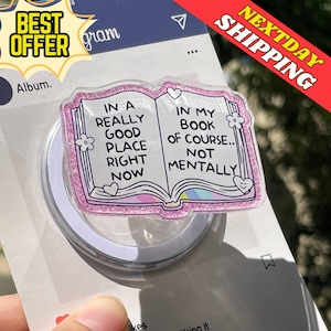 Puede incluir: Un soporte transparente para teléfono con un borde de purpurina rosa, con un diseño de libro con el texto "IN A REALLY GOOD PLACE RIGHT NOW" y "IN MY BOOK OF COURSE.. NOT MENTALLY". El soporte tiene una base circular blanca.