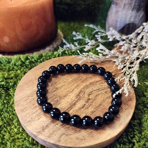 May include: A black beaded bracelet sits on a small, round wooden coaster. The bracelet is made of polished, round black beads. A lit candle and dried flowers are in the background, set against a green, textured surface.
