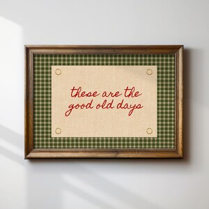 以下が含まれることがあります： 赤い筆記体で「these are the good old days」と書かれた額入りのプリント。プリントは、緑と白のチェック柄のボーダーと濃い茶色の木製フレームが付いたタン色の背景です。プリントは白い壁に掛けられています。