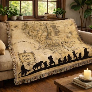 May include: A beige throw blanket with a map of Middle Earth, featuring black silhouettes of characters and the text "Middle Earth." The blanket has a fringed edge and is draped over a sofa.