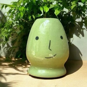 May include: A light green ceramic vase with a whimsical face design. The vase has a rounded body and a small base. Black dots and a line create the eyes, nose, and mouth. Green foliage is visible in the background.