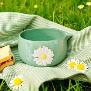 Puede incluir: Un cuenco de cerámica verde claro con una aplicación de margarita blanca, sobre un paño a rayas verdes y blancas. También hay un sándwich y varias margaritas, en una escena al aire libre con hierba.