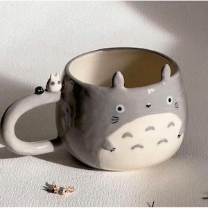 May include: A ceramic mug shaped like a Totoro character, with gray and white coloring. The mug has a handle with a small, white cat-like figure and a black dot. The mug features Totoro's facial features and ears.