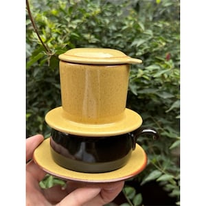 May include: A yellow ceramic Vietnamese coffee filter set, comprising a filter, lid, and cup with a handle, resting on a saucer. The background features green foliage.