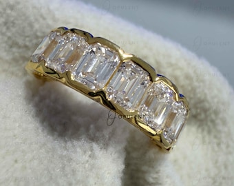 May include: A gold band ring featuring a row of five clear emerald-cut stones set in a shared prong setting. The metal has a polished yellow finish, and the stones are arranged horizontally across the top of the band, creating a continuous line.